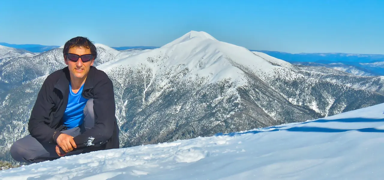 Chris Rishworth in sunglasses crouching in the snow with Mt Feathertop behind