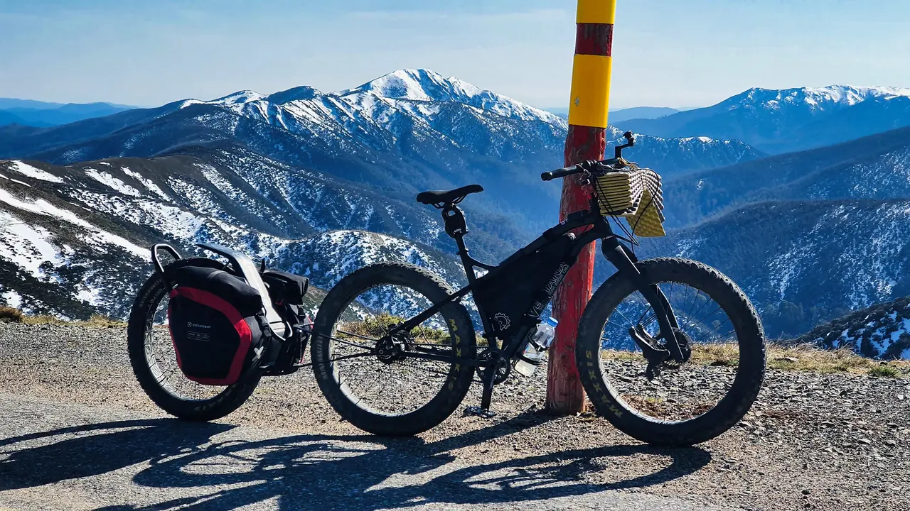 Fat bike and a one-wheel trailer with red and black panniers, leaning on a snow pole with a snow-capped peak behind