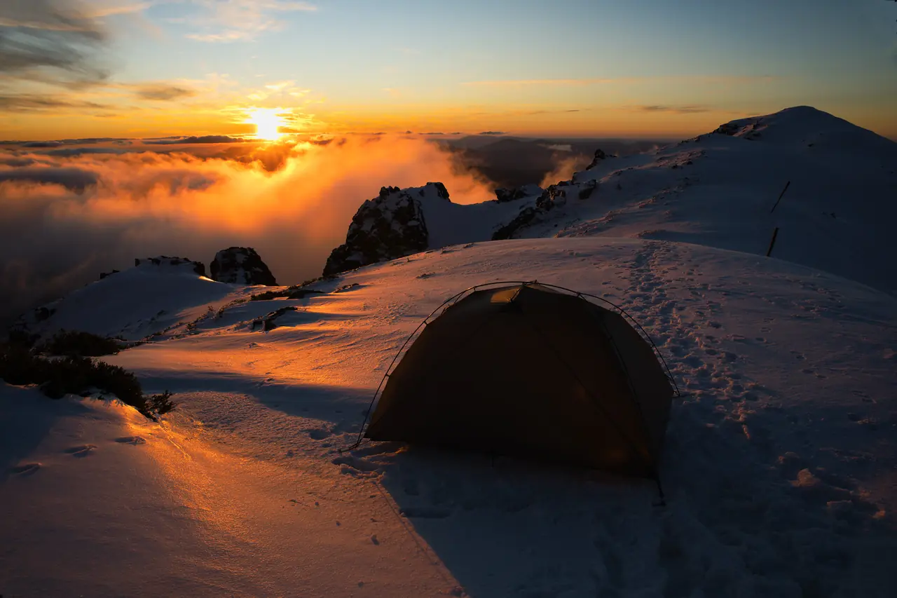 Snow-covered tent glowing at sunset above a sea of cloud