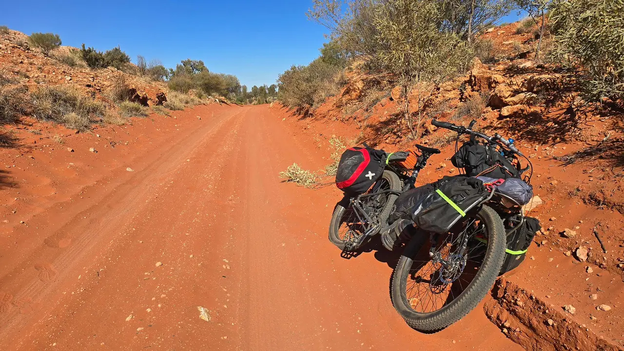 Black fat bike with frame bag and rear panniers leaning on a red dirt track lined with desert scrub