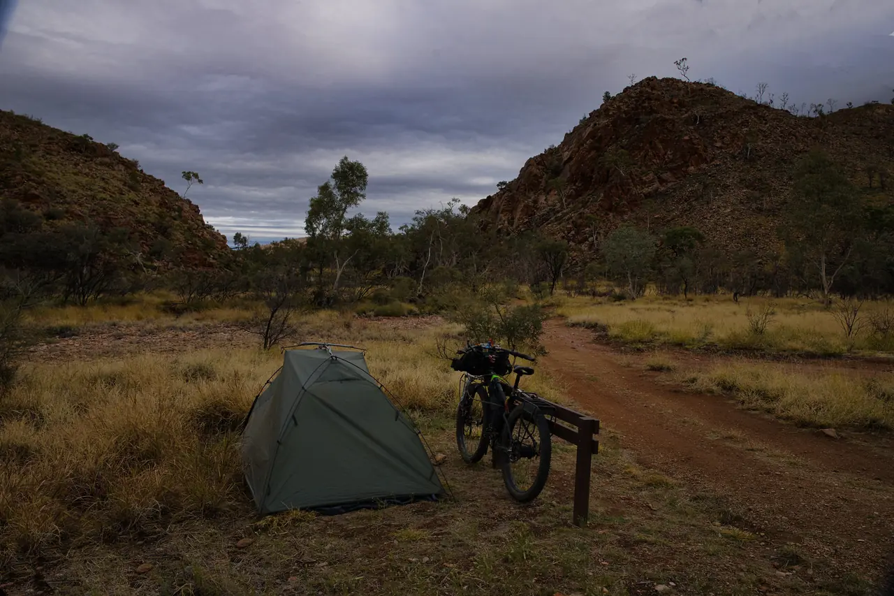 Small green tent and fat bike at a bush camp under rocky red hills and a heavy grey sky