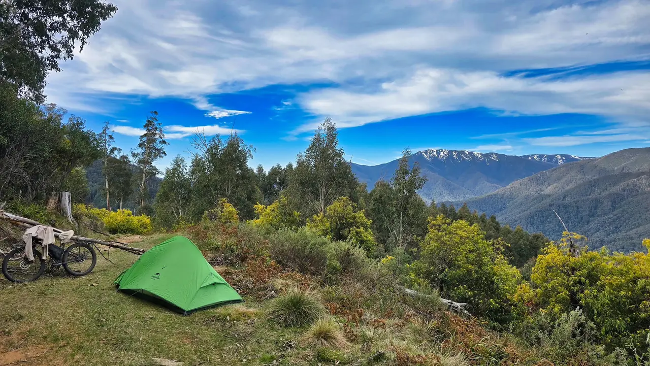Green tent and fat bike on a grassy ridge with snow on the distant ranges